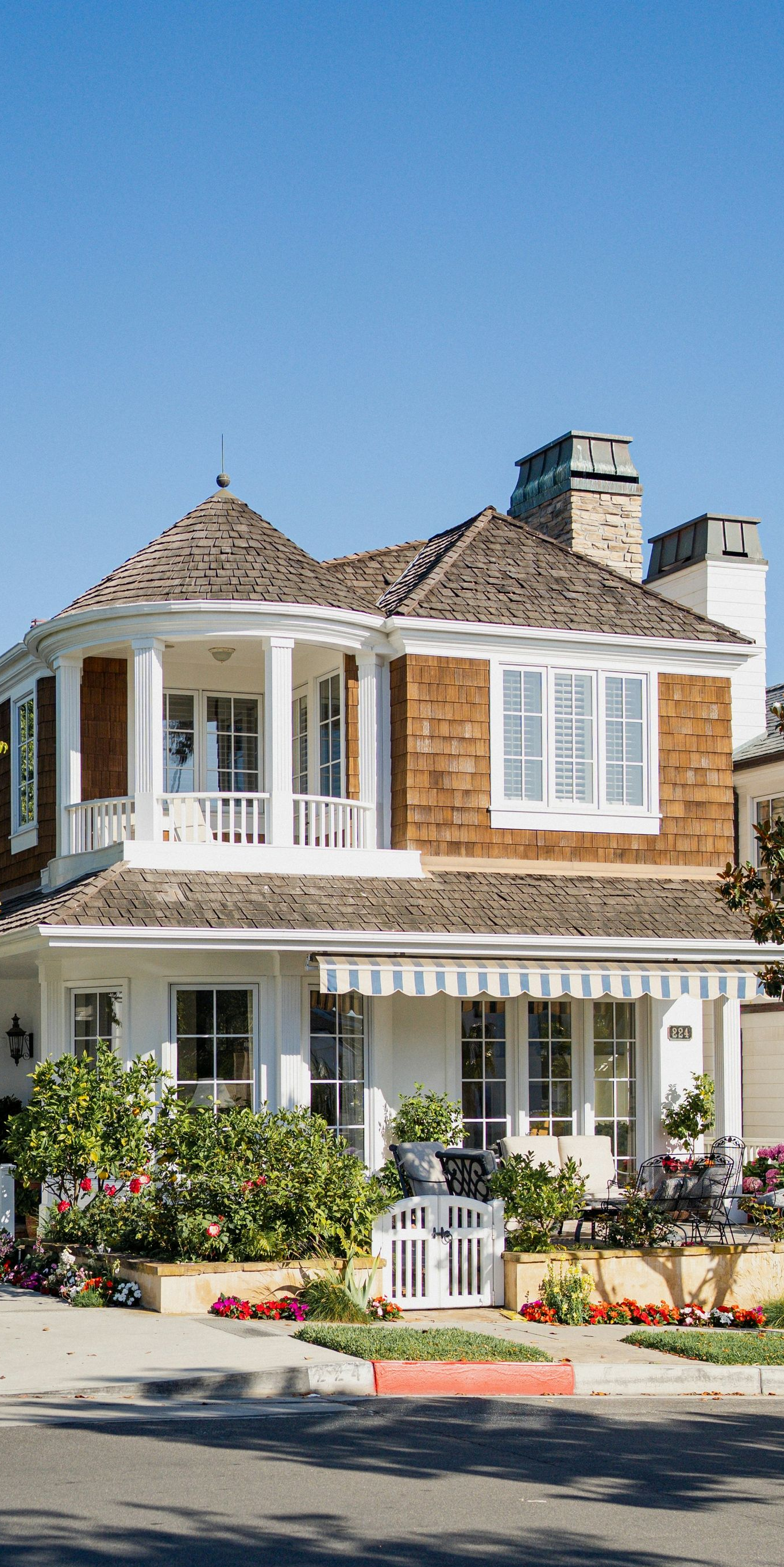 Two-story white house with brown shingles, blue sky. Green bushes and red flowers in front.