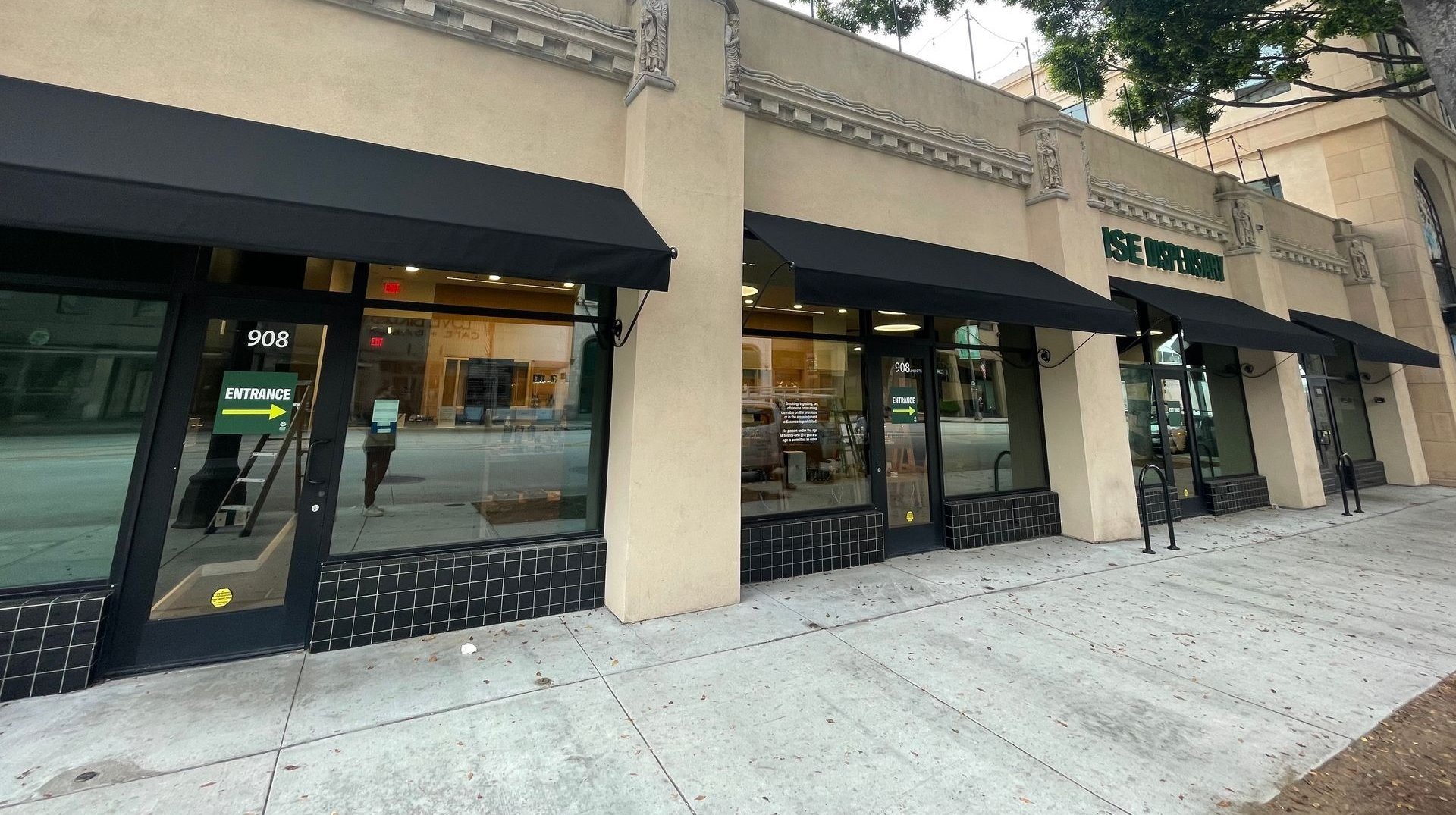 Exterior of a beige building with black awnings and large windows. People walking on the sidewalk.