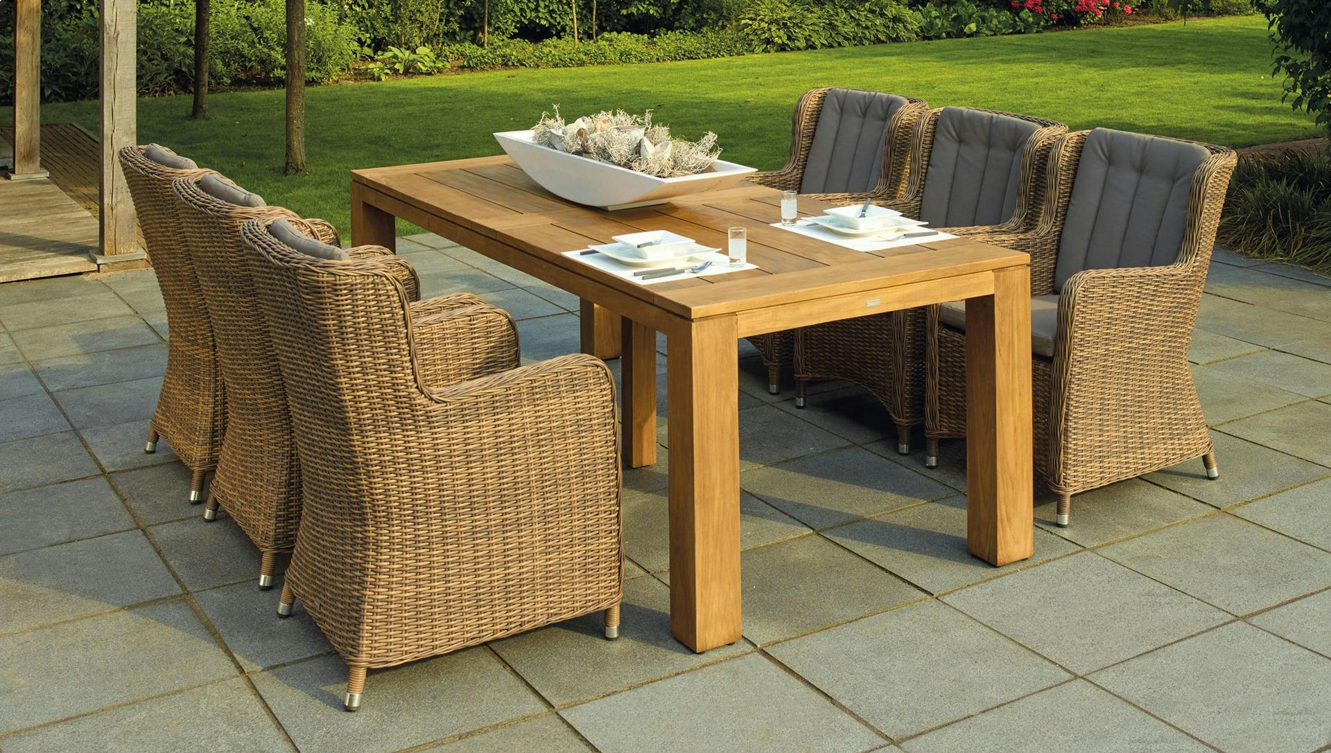 Outdoor dining set: wooden table with six woven chairs on a stone patio, with a garden background.