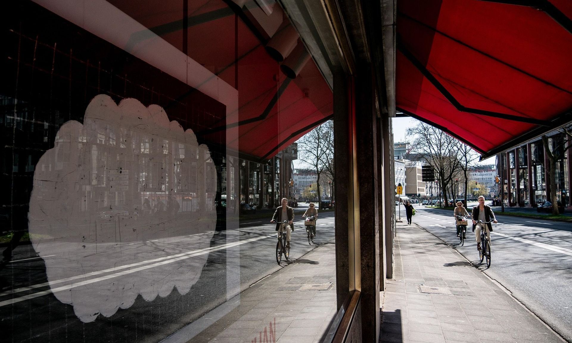A storefront reflects the street with people on bikes. Red awning above. White cloudy shape on the storefront.