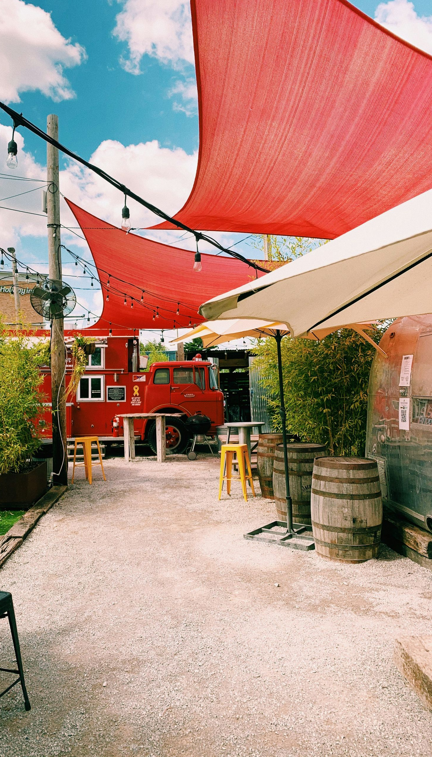 Outdoor patio with red sun shades, umbrellas, and a red food truck, gravel ground.
