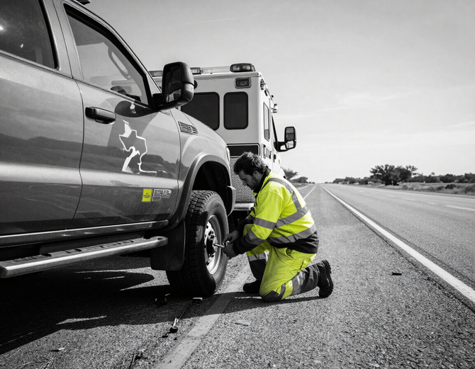 Roadside assistance technician changing a flat tire beside an emergency vehicle.