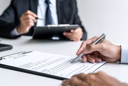 A person signing a document on a clipboard, while another person in a suit holds a pen and notepad.