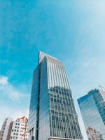 A modern skyscraper with a pointed top, reflecting the blue sky, next to a smaller glass building.