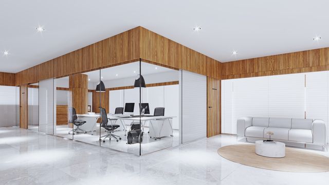 Modern office space with glass-walled offices, wood paneling, and a white sofa in the lounge area. The floor is white, and there are computers visible.