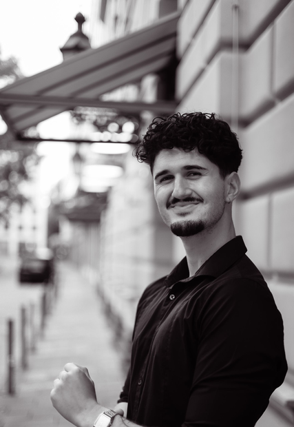 Man in a black shirt smiling and flexing his arm on a city sidewalk under an awning