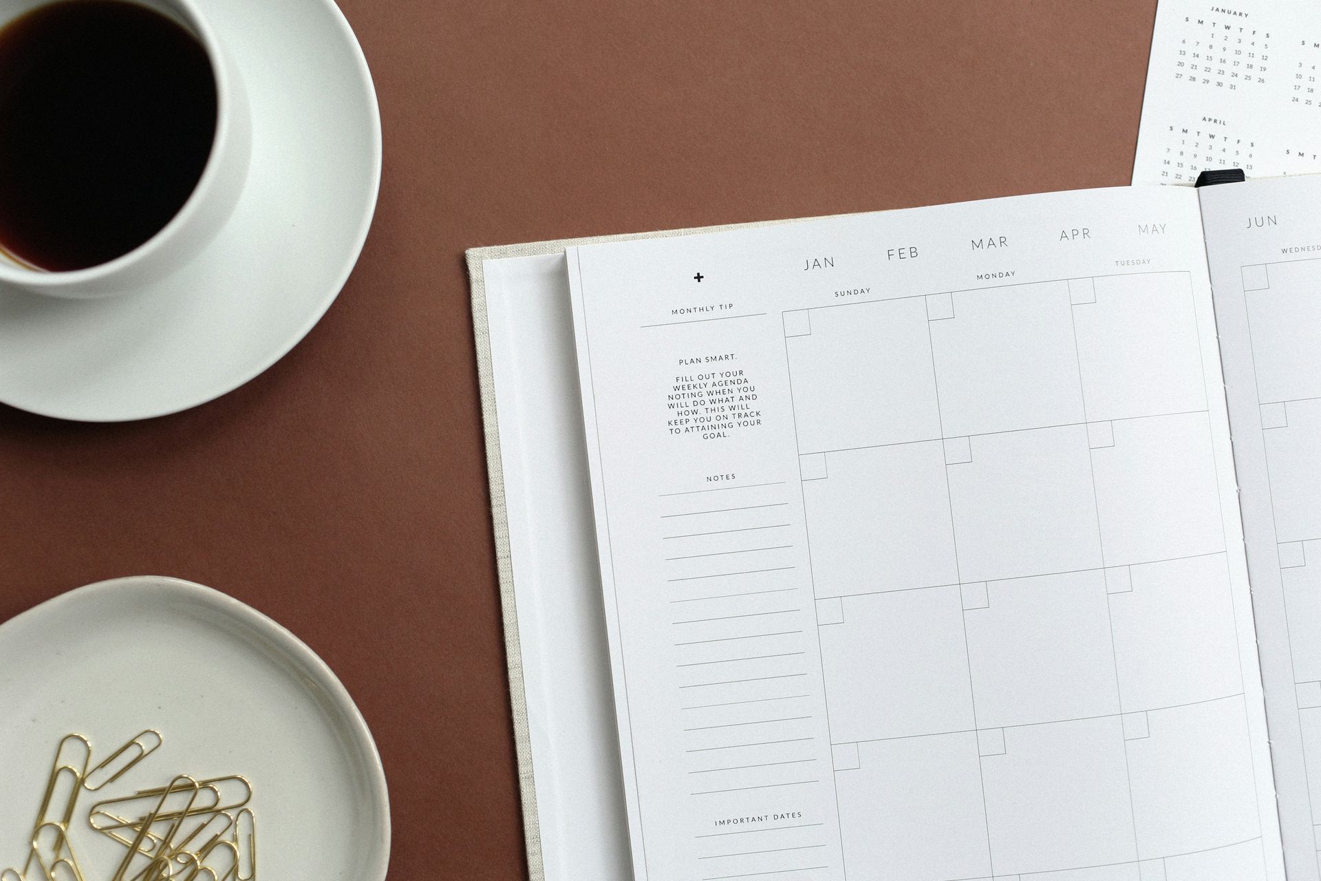 Eine Tasse Kaffee, ein Teller mit Büroklammern und ein Kalender auf einem Tisch.