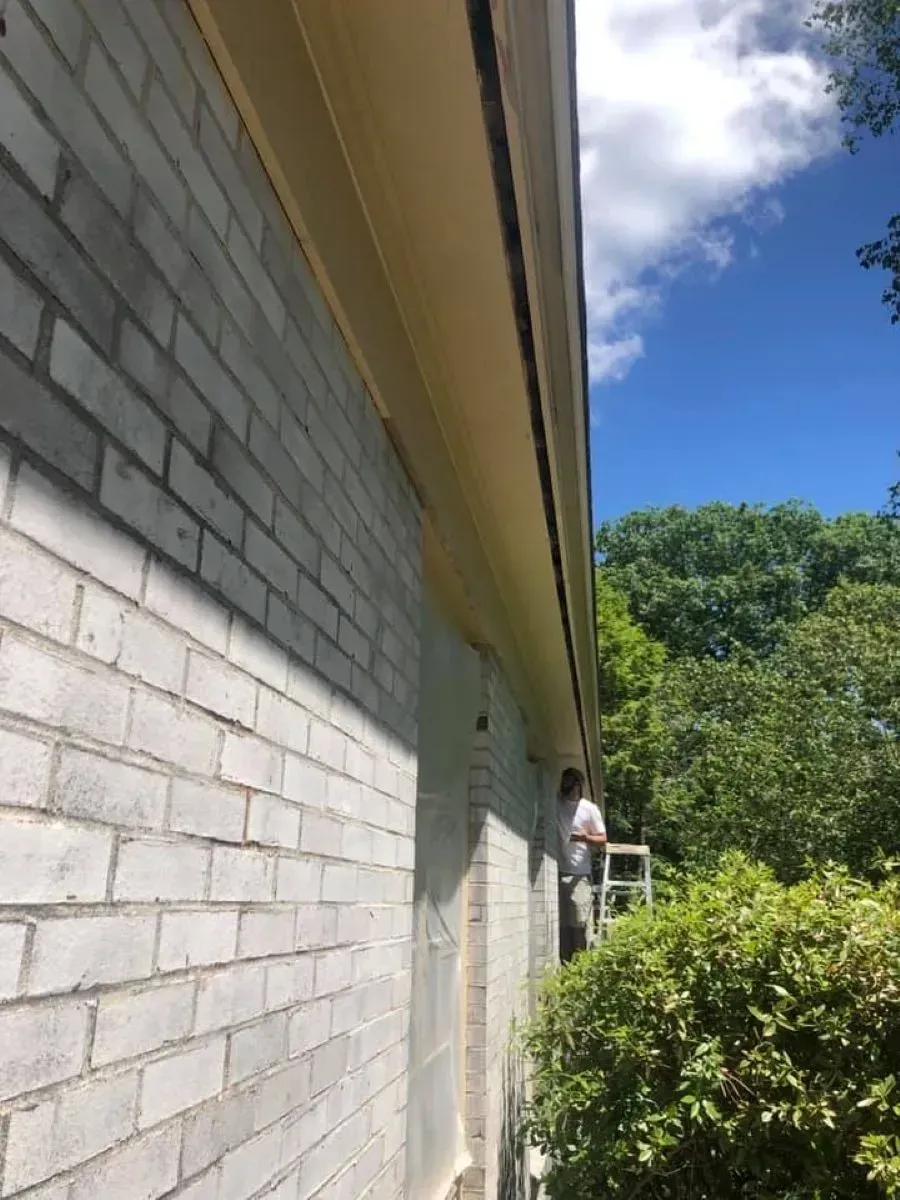 A man is standing on a ladder painting the side of a white brick house.