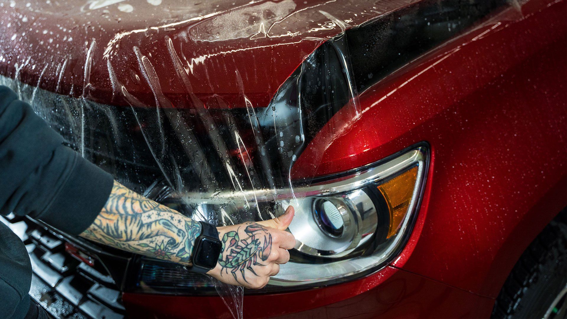 A man with tattoos is covering the hood of a red car with plastic.