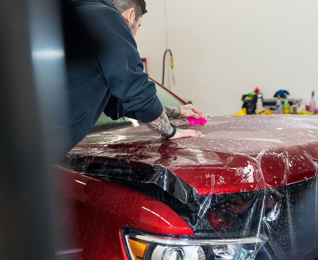 A man is covering the hood of a red car with plastic wrap.