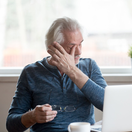 Older man, rubbing eye, sits at a laptop. Holds glasses, wearing blue shirt, near a window.