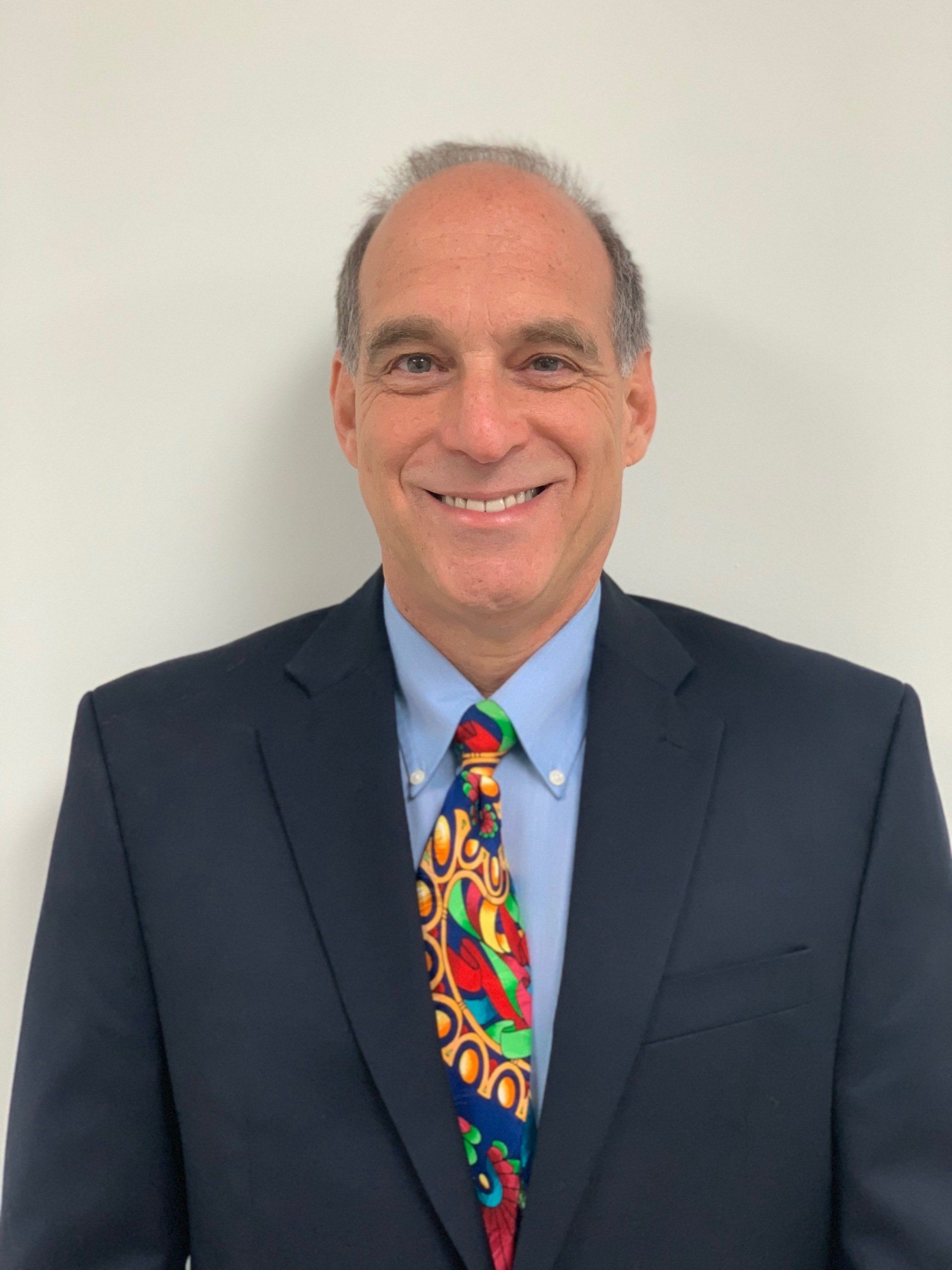 Smiling man in a suit and tie, standing against a plain white wall.