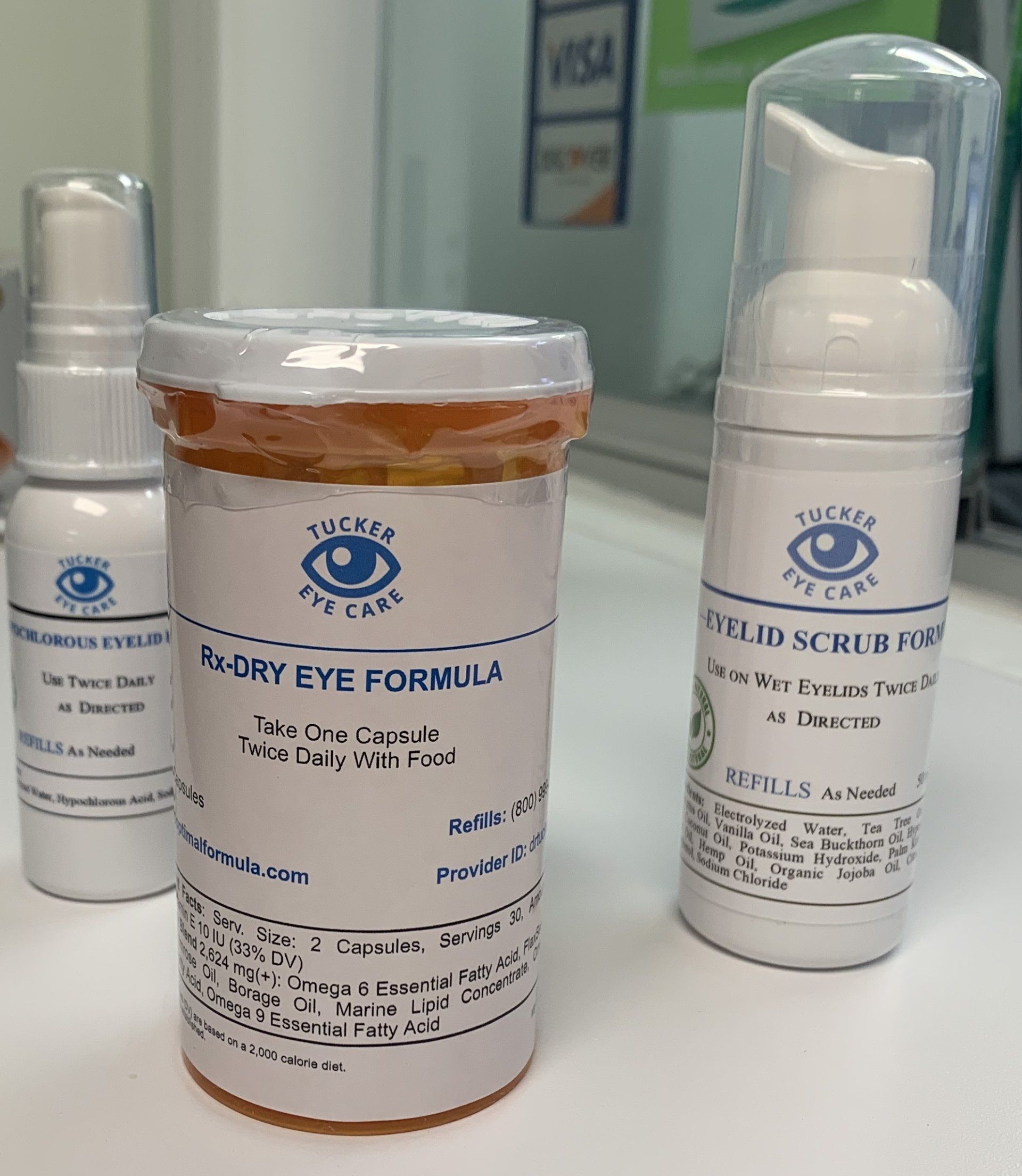 Three eye care products on a counter: tear spray, dry eye formula in a pill bottle, and eyelid scrub foam.
