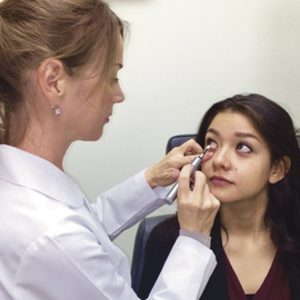 Optometrist examining a young woman's eye with a light in a clinic.