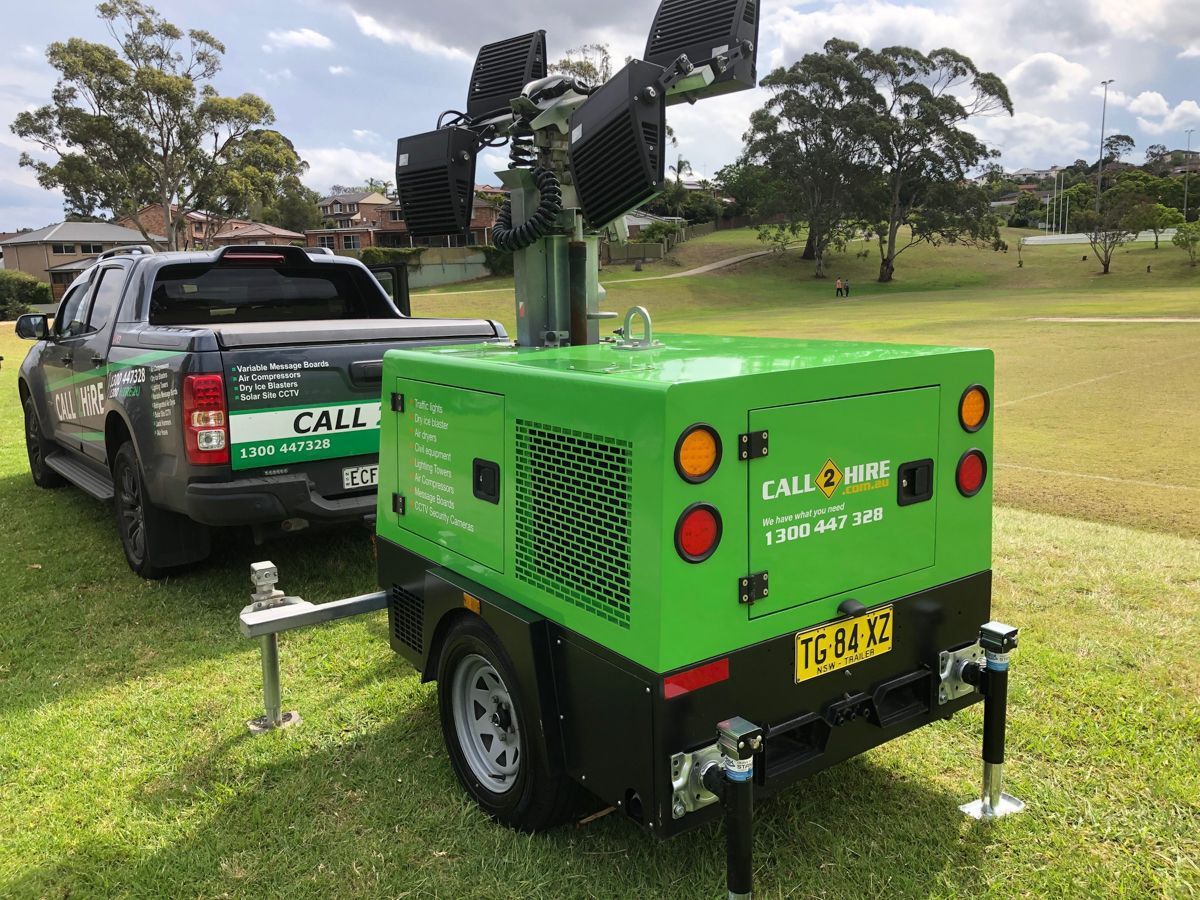 A green portable light tower trailer is parked on a grassy field, attached to a pickup truck.