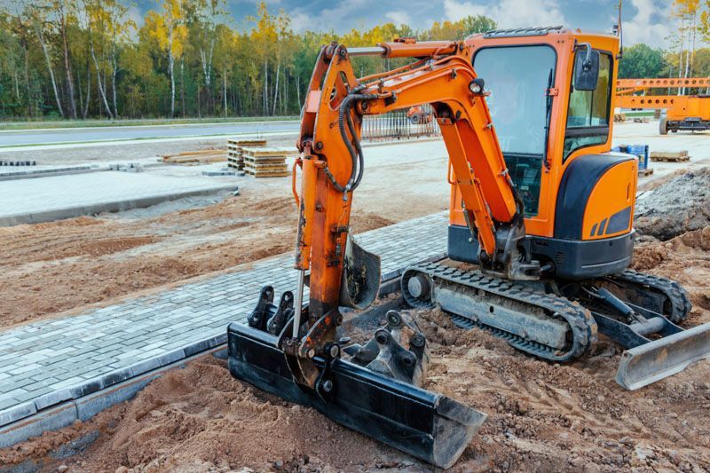 An orange mini excavator stands on a construction site next to a newly paved stone walkway.