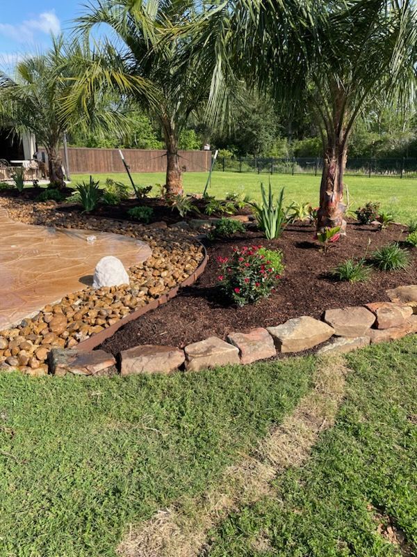 A stone-edged garden bed with mulch, small plants, and palm trees curves around a patio area with a stone rock feature.