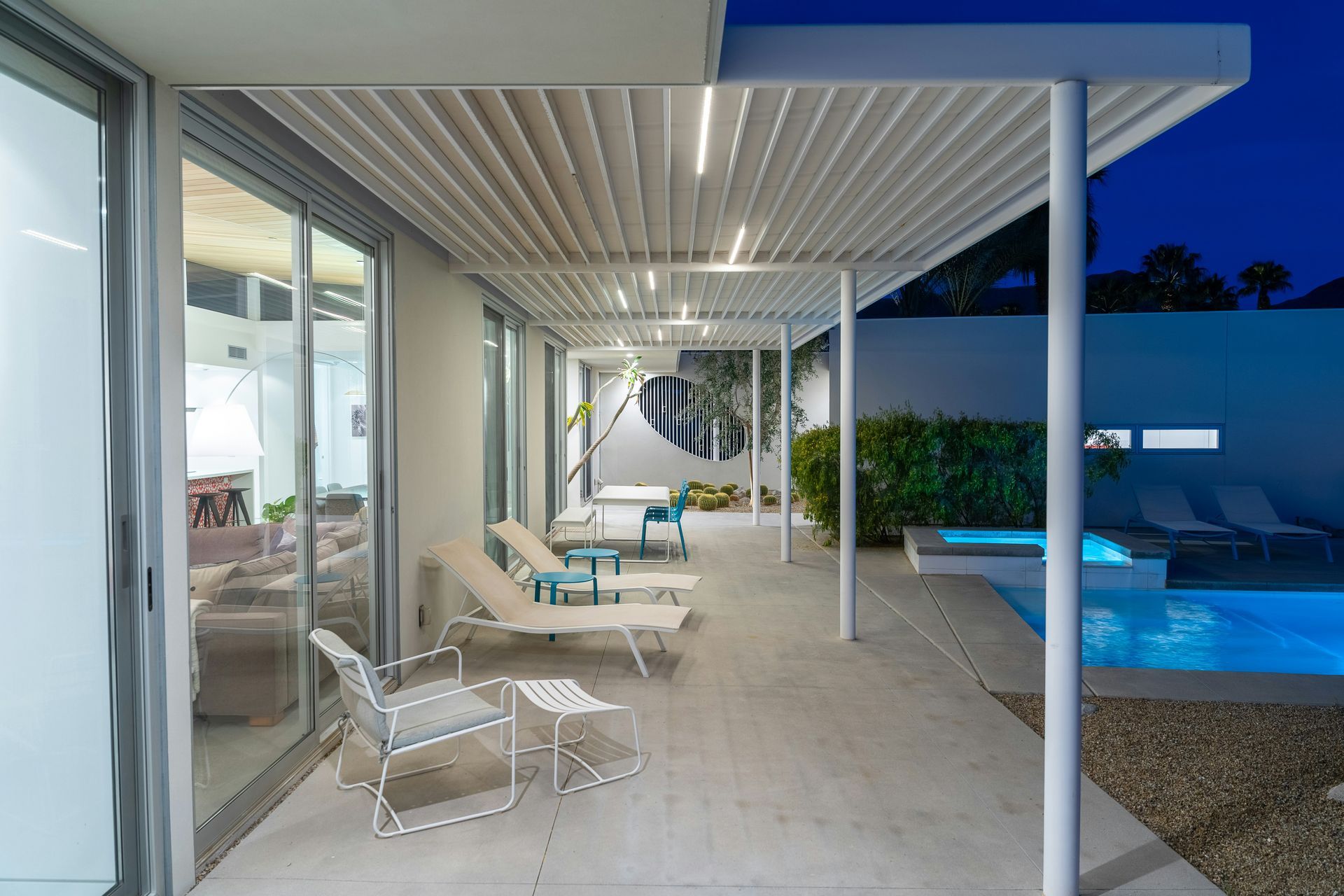 Modern patio with white lounge chairs, covered ceiling, and a lit blue swimming pool at dusk