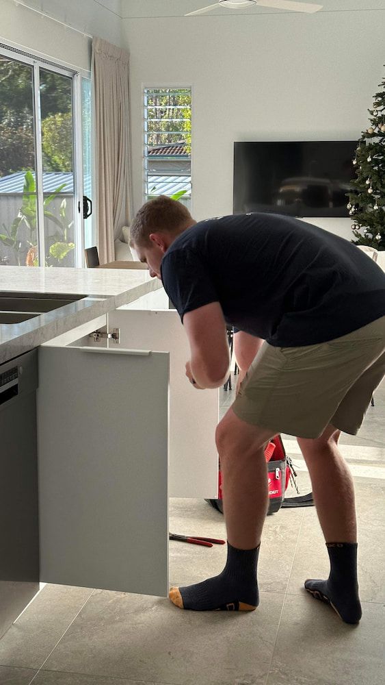 A Man is Kneeling Down in a Kitchen Looking Into a Cabinet — R.K. Plumbing & Gas In Bli Bli, QLD