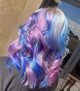 A woman with purple and blue hair is standing in front of a window.