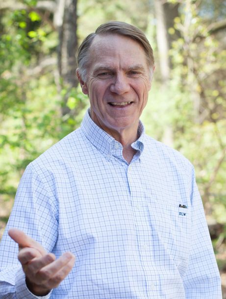 A man in a white shirt is standing in the woods and smiling.