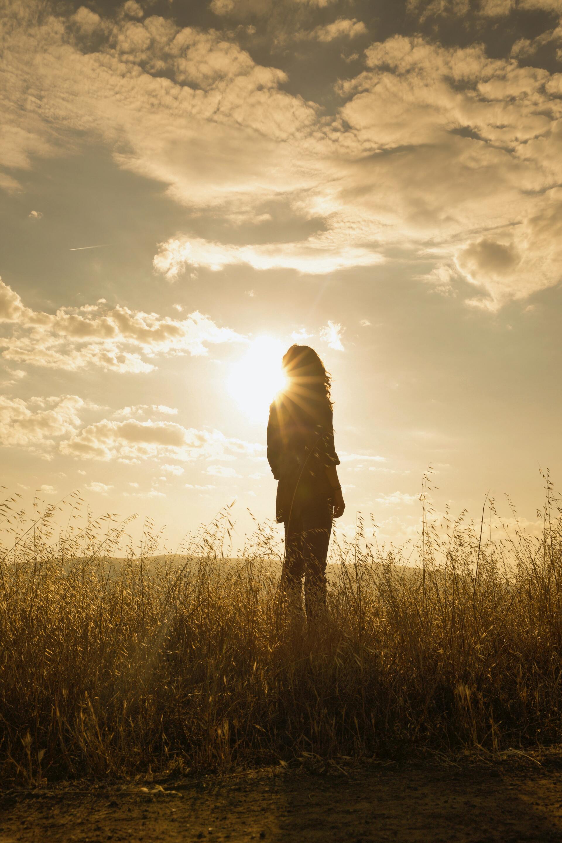 A woman is standing in a field at sunset looking at the sun.