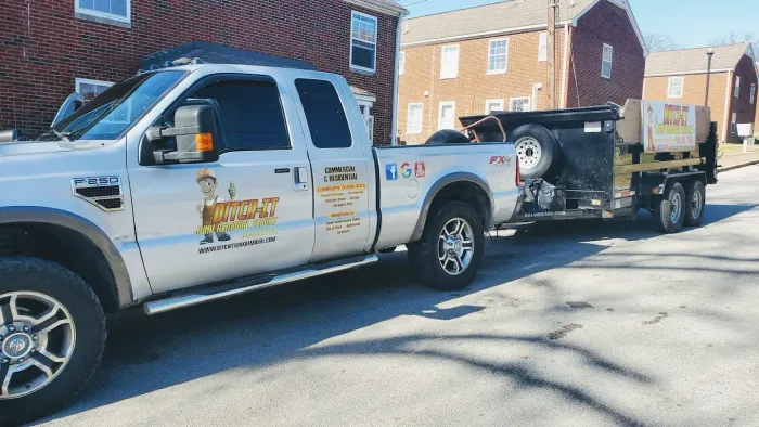 Silver pickup truck towing a trailer with equipment, parked on a street near brick buildings.