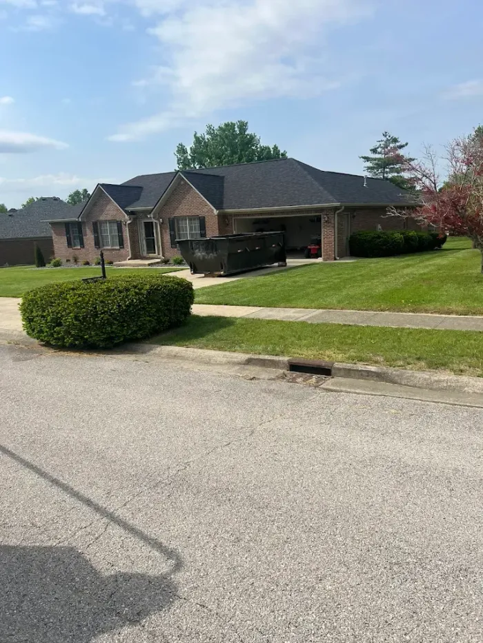 Brick house with dark roof and green lawn; trash dumpster in driveway on sunny day.