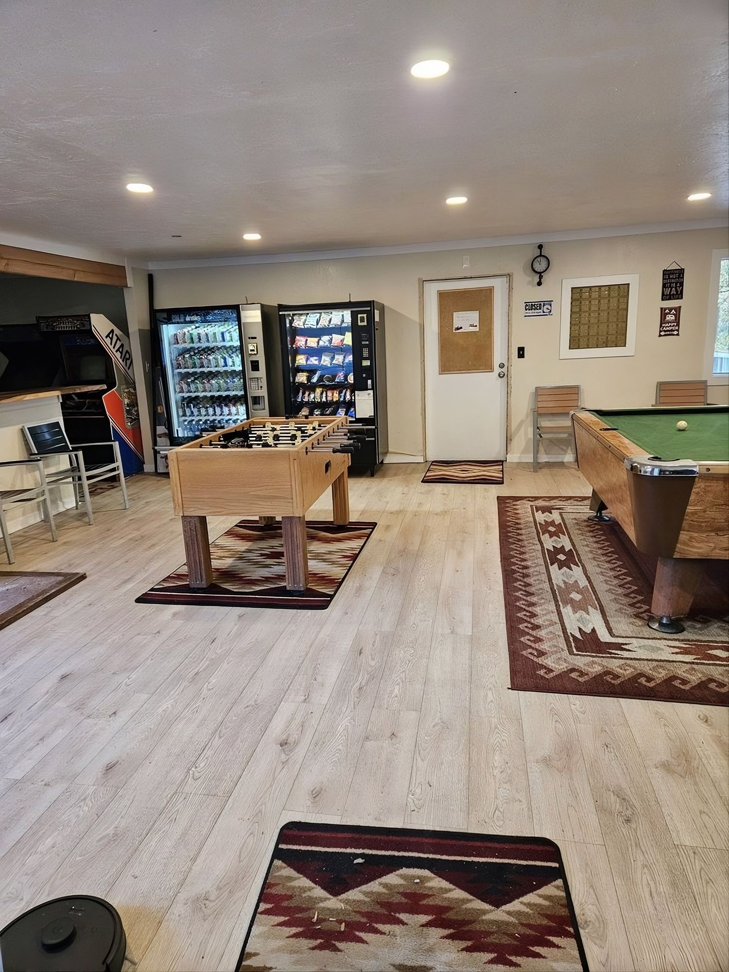 Game room with foosball, pool table, vending machines, rugs on light wood floor.