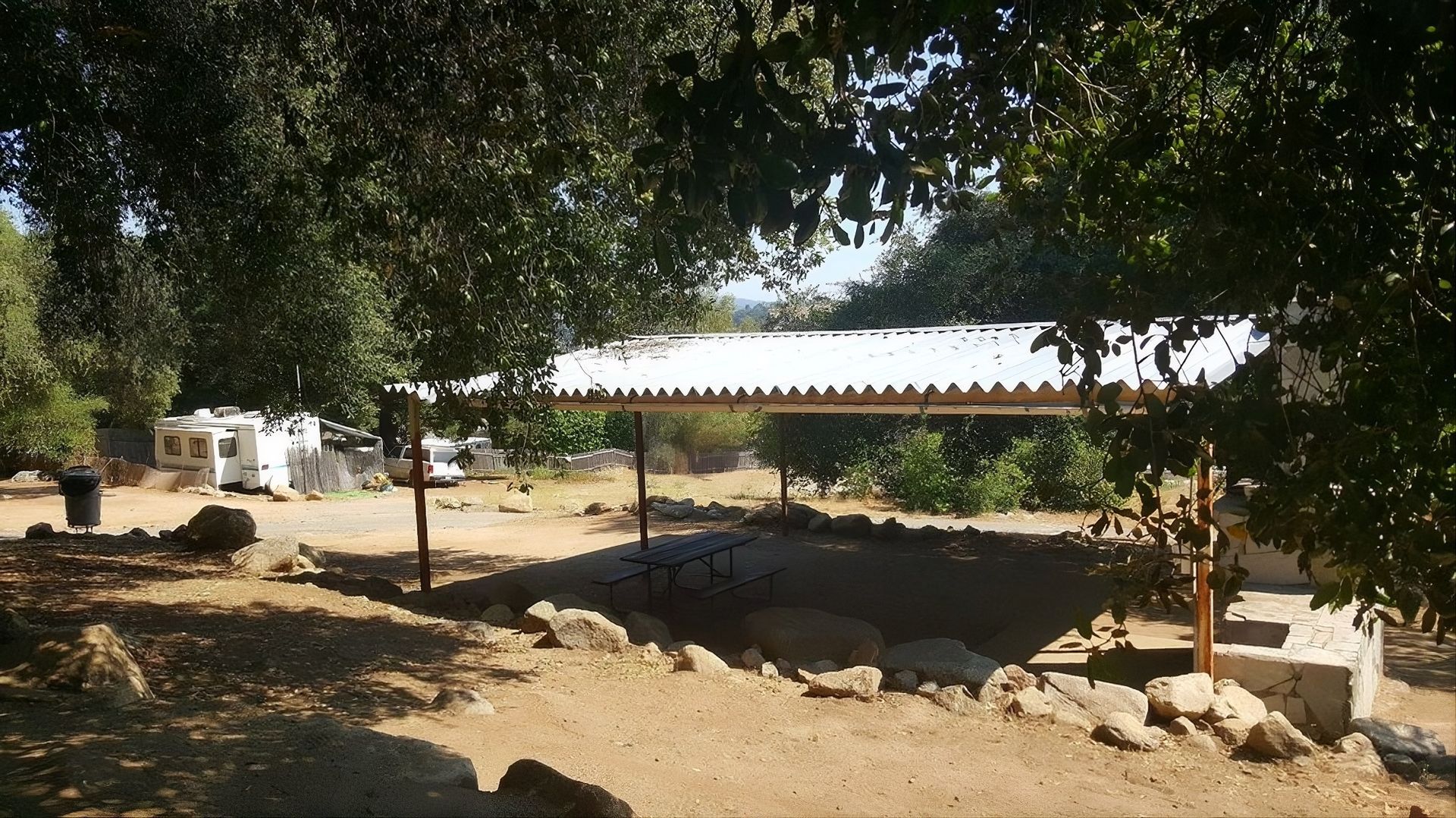 A covered structure with a stone perimeter in a sunny outdoor setting with trees.
