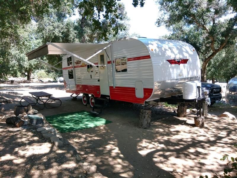 White and red vintage camper with awning extended, parked in a shaded campsite near a picnic table.