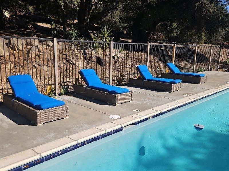Four blue-cushioned lounge chairs by a swimming pool, fence in the background, sunny day.