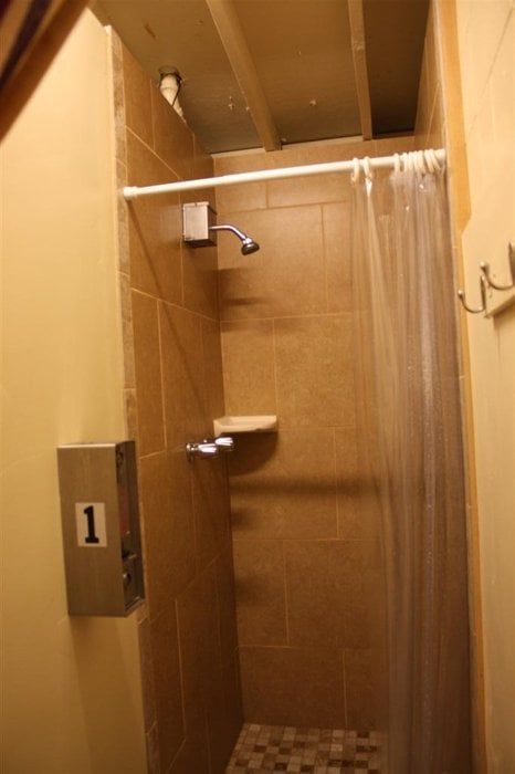 Shower stall with tan tiled walls, showerhead, and a clear shower curtain. Soap dispenser on the wall.