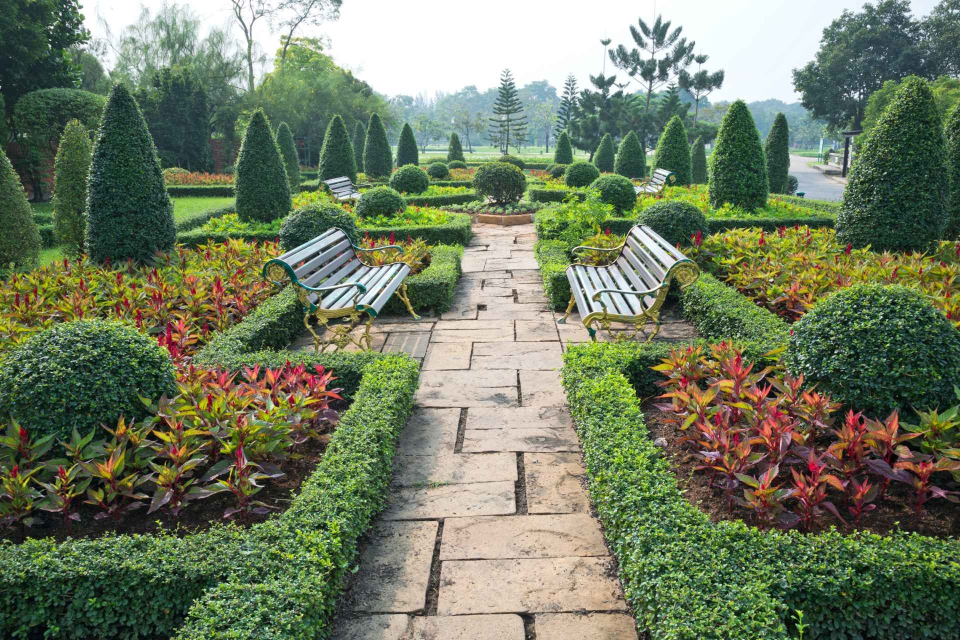 Stone path through a formal garden with manicured bushes, trees, and benches, with colorful foliage in the beds.