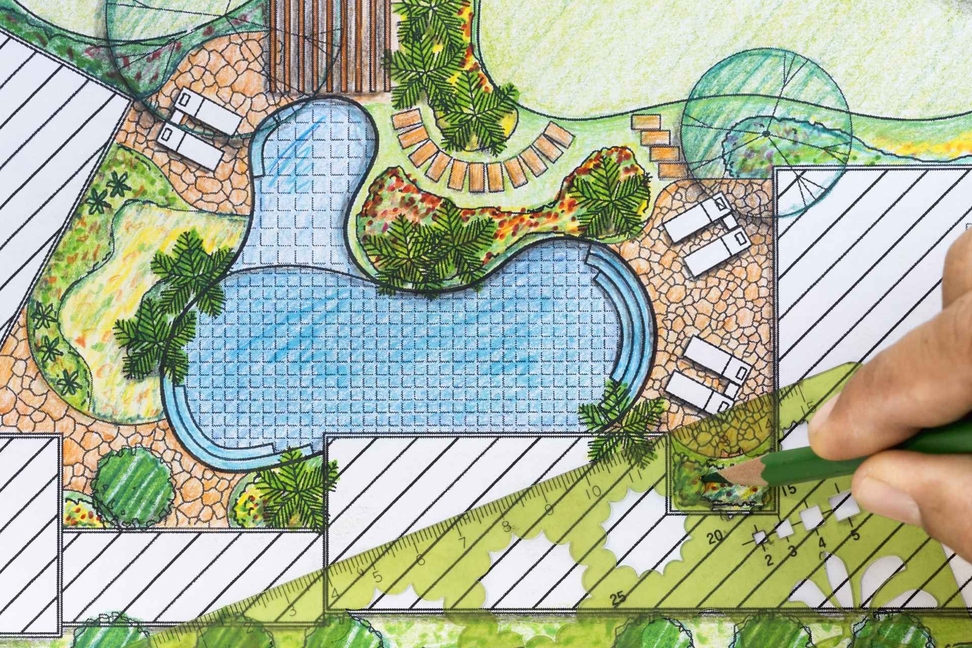 Hand coloring a landscape design plan with a green pencil, showing a pool, trees, lounge chairs, and paved areas.