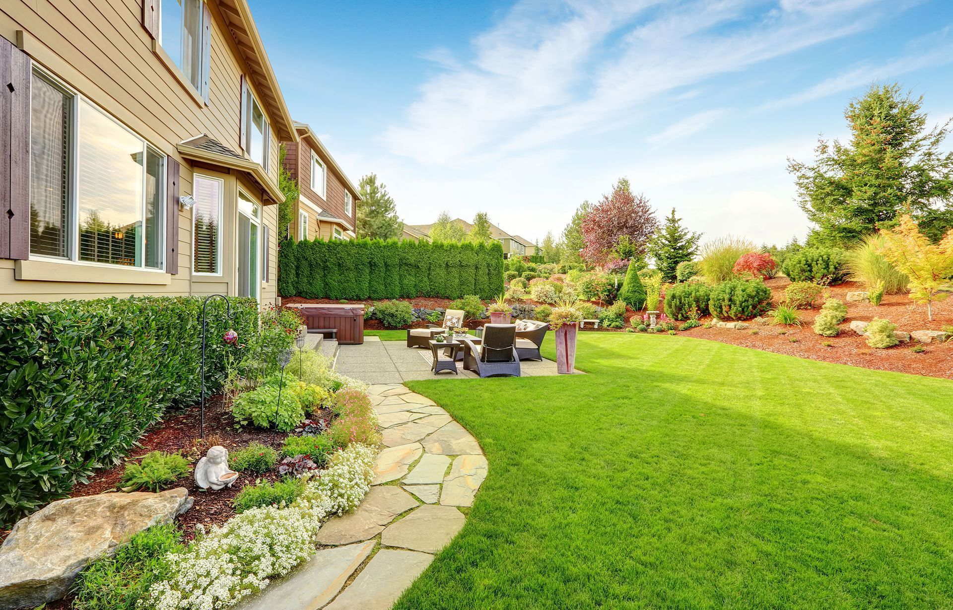 Lush backyard with stone path, green lawn, and flowerbeds, leading to a patio with seating. The house is visible on the left, under a bright blue sky.