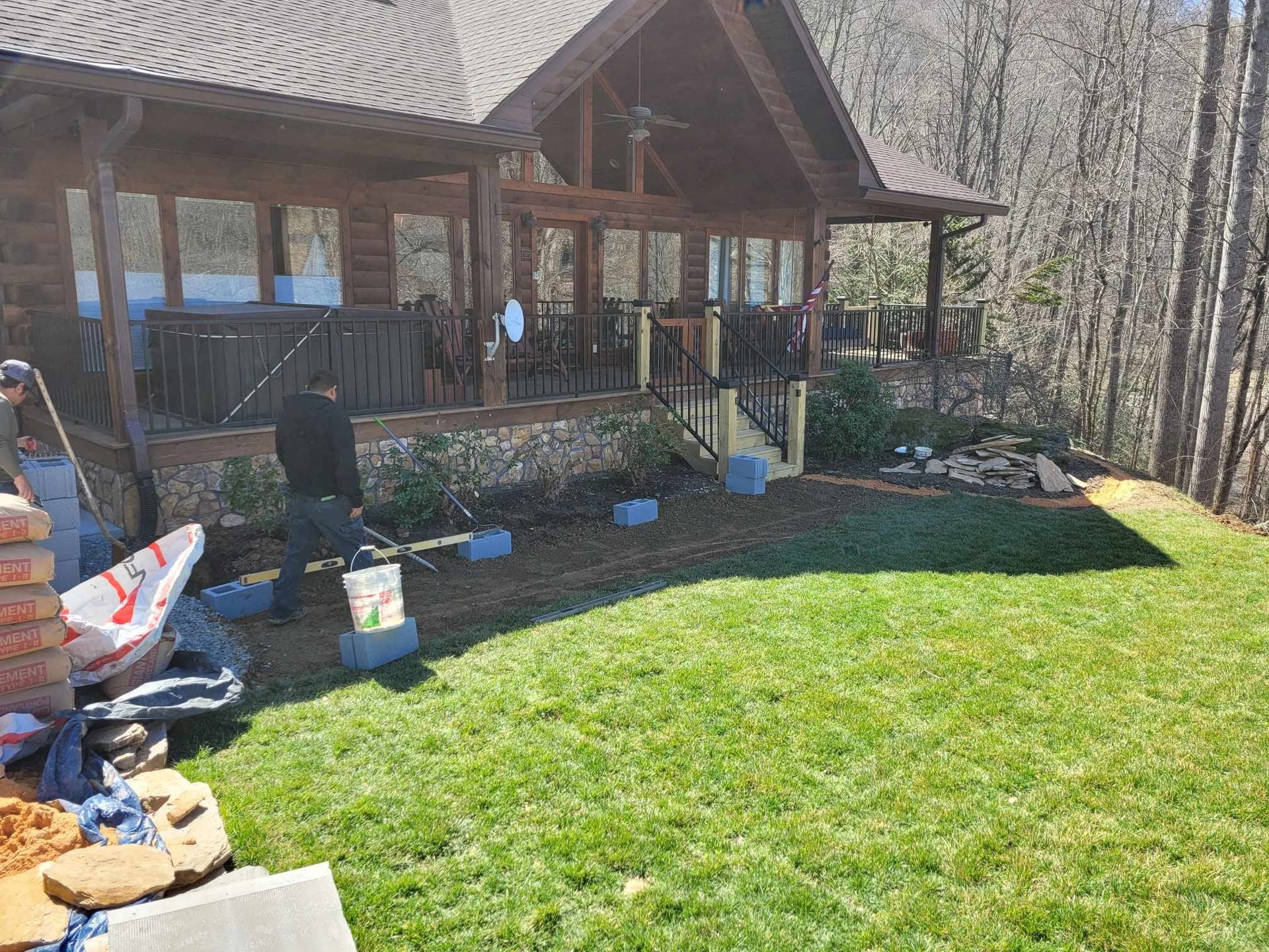 A person is working on a landscaping project near a log cabin with a porch. They are in a grassy area with materials nearby.