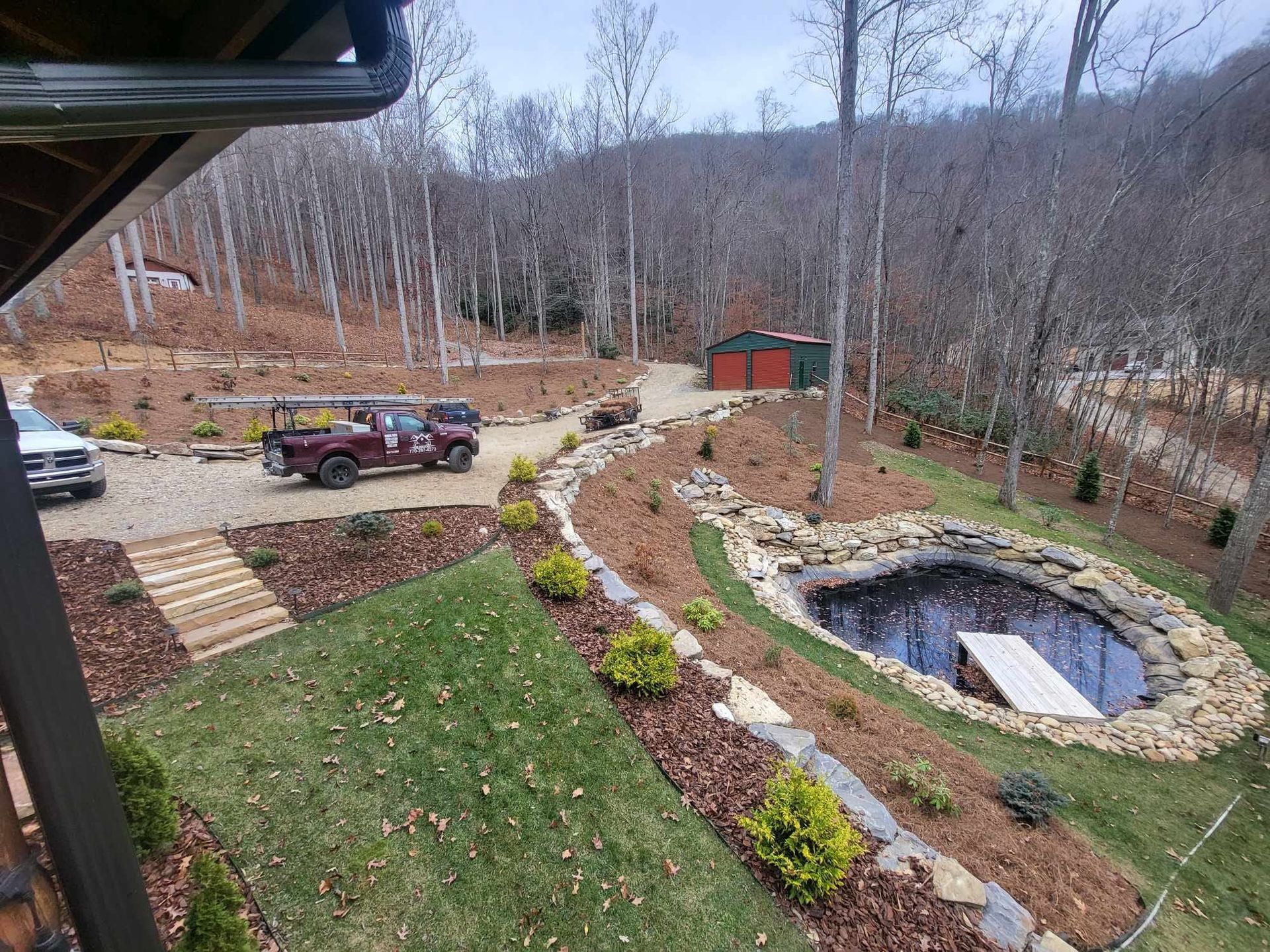 A mountain landscape with a pond, landscaping, and a red truck parked on a gravel driveway. A building can be seen further back.