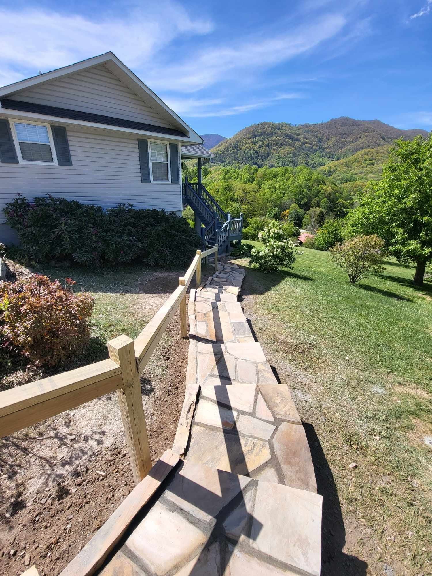 Stone steps and wooden fence leading up to a light-colored house with a mountain backdrop and blue sky.