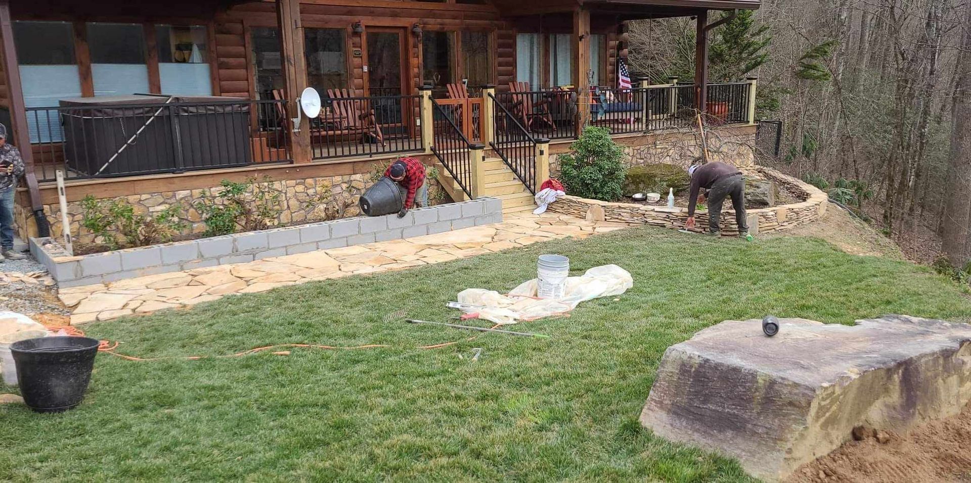 Workers landscaping the yard of a log cabin. One person places pavers, while others work on a retaining wall.