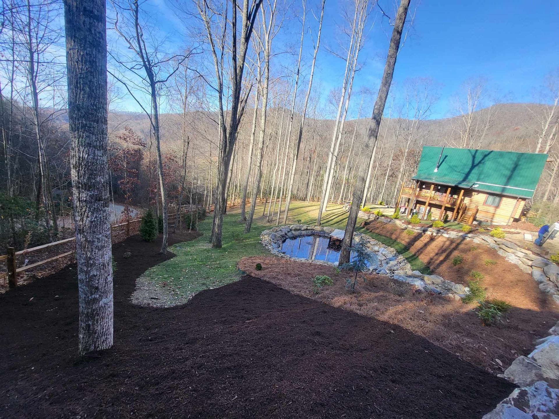 Cabin with a green roof nestled among trees near a small pond, on a hillside.