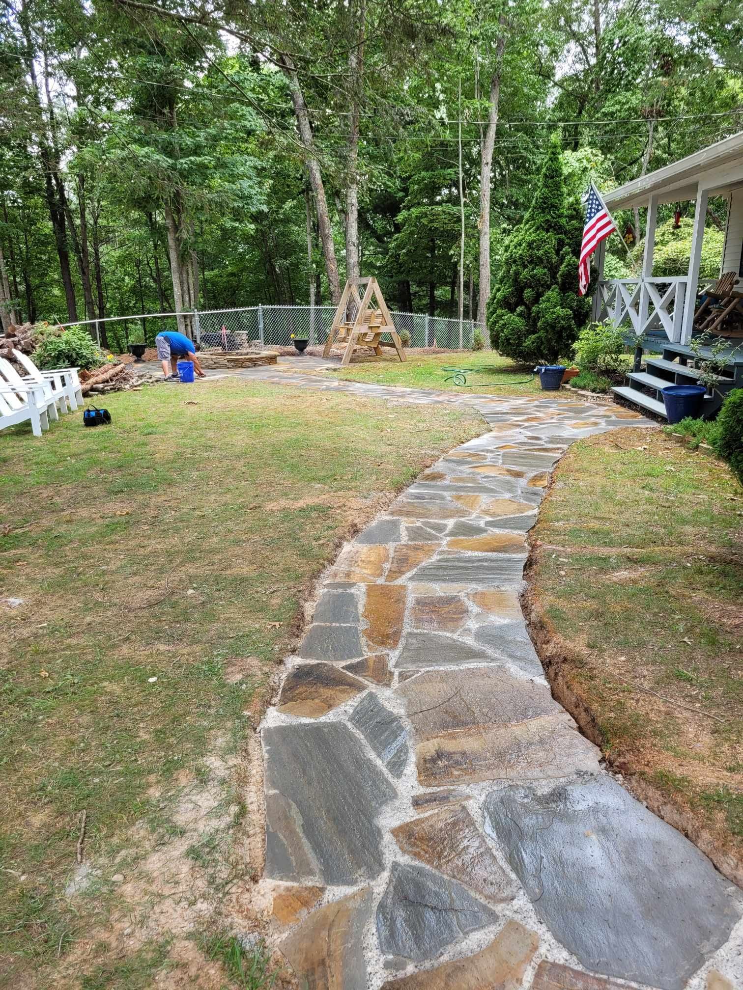 Stone pathway leading through a yard towards a house with a porch and an American flag. A person is working in the background.