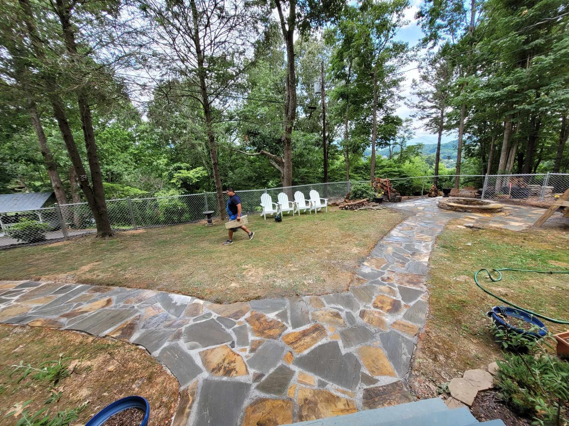 A person walks across a grassy backyard with a stone path. White chairs sit in the distance. Trees and a fence surround the yard.