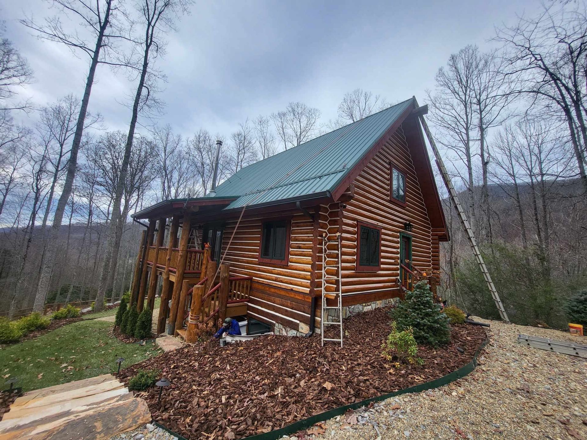 A brown log cabin with a green roof stands in a wooded area under a cloudy sky. There is a ladder next to the cabin.