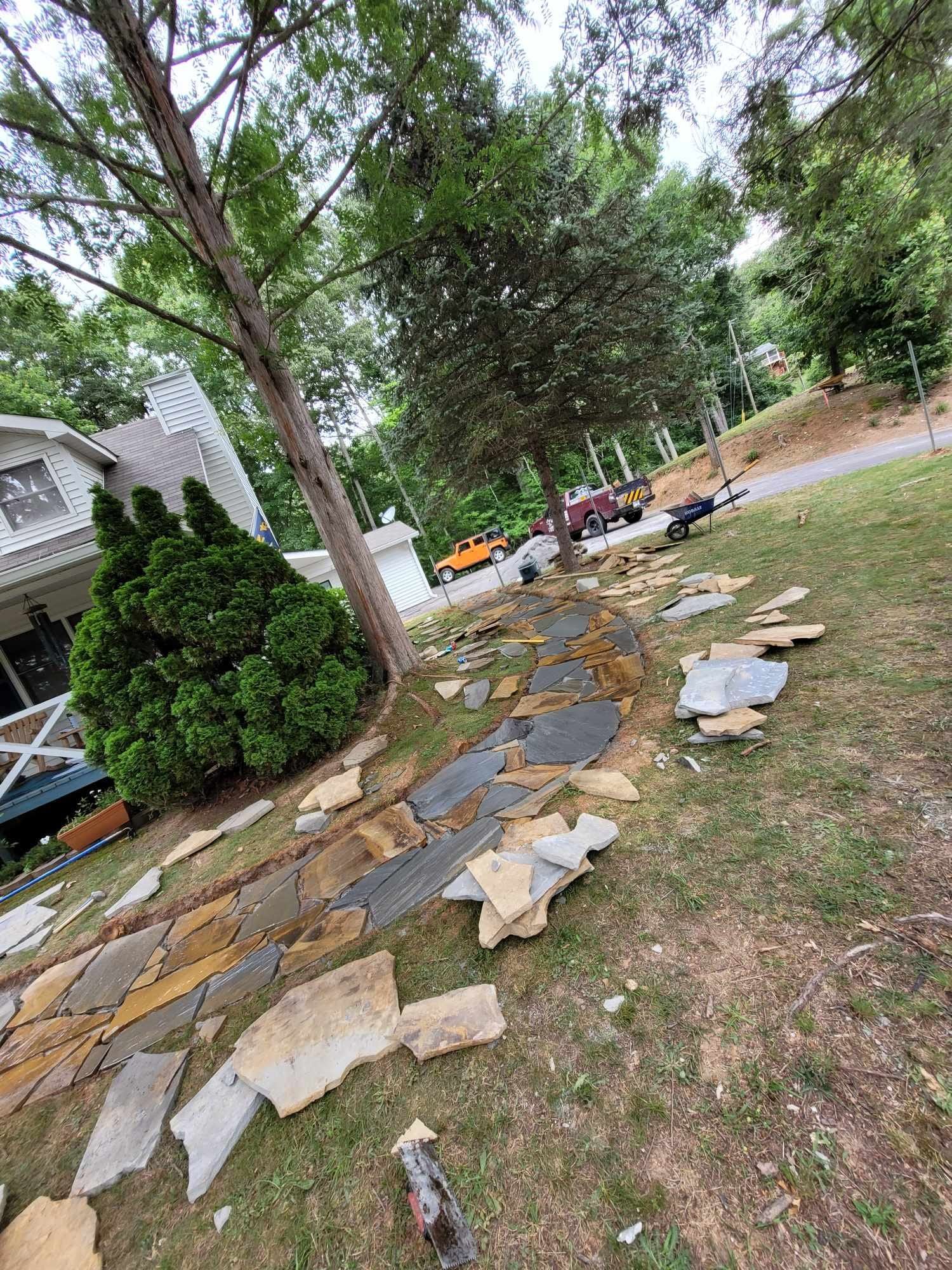 A pathway under construction with flat stones of varying colors, leading toward a road lined with trees.