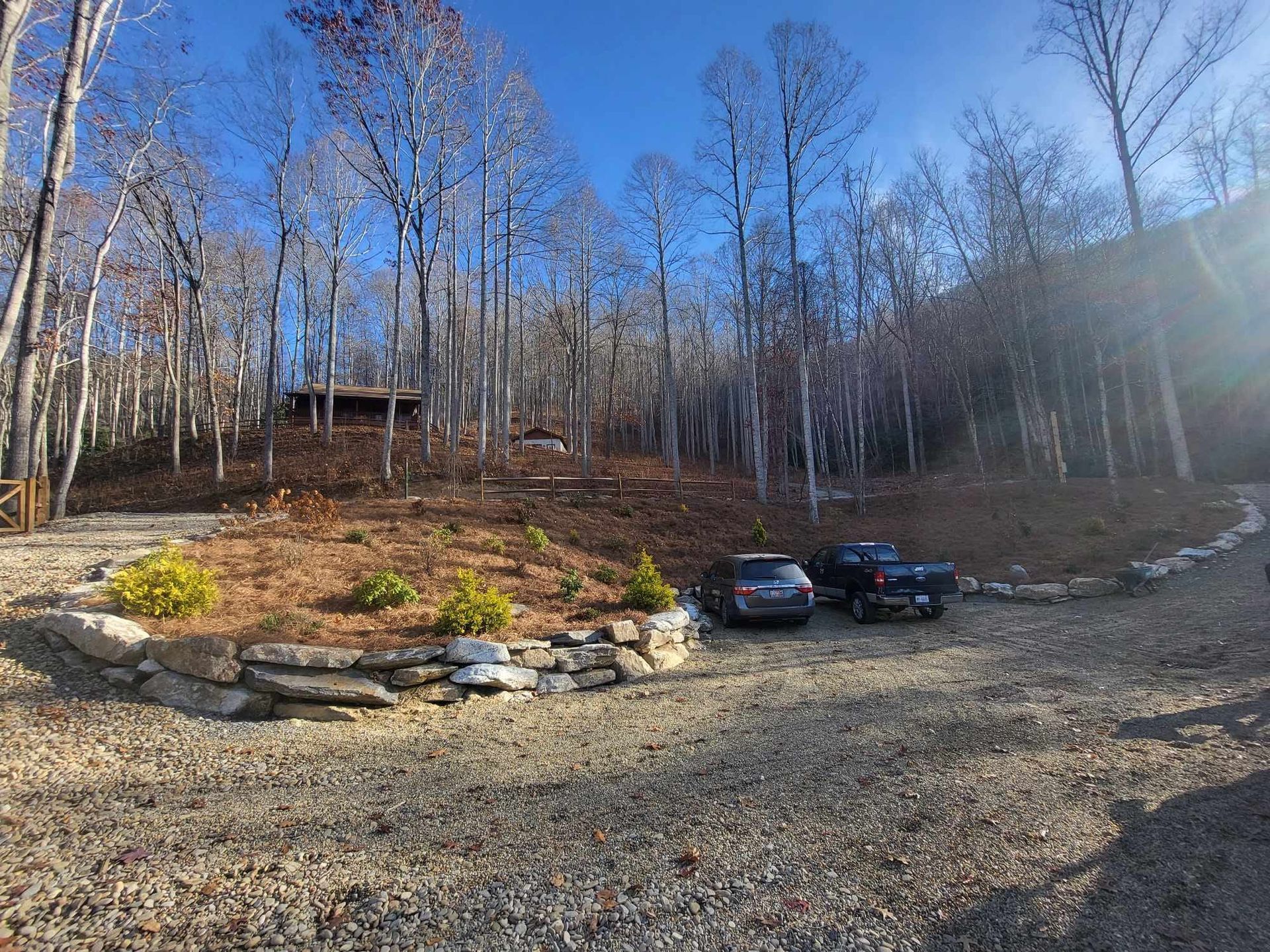 Gravel driveway leads to a cabin in the woods with a few parked cars, surrounded by bare trees under a blue sky.