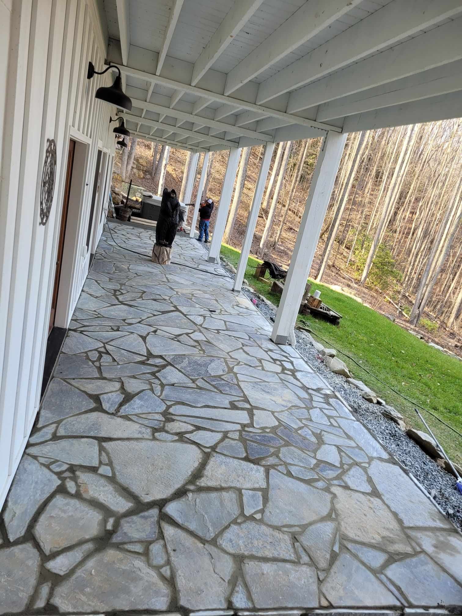A long, covered porch with irregular stone flooring.  People stand near the middle, and a forest lines the edge.