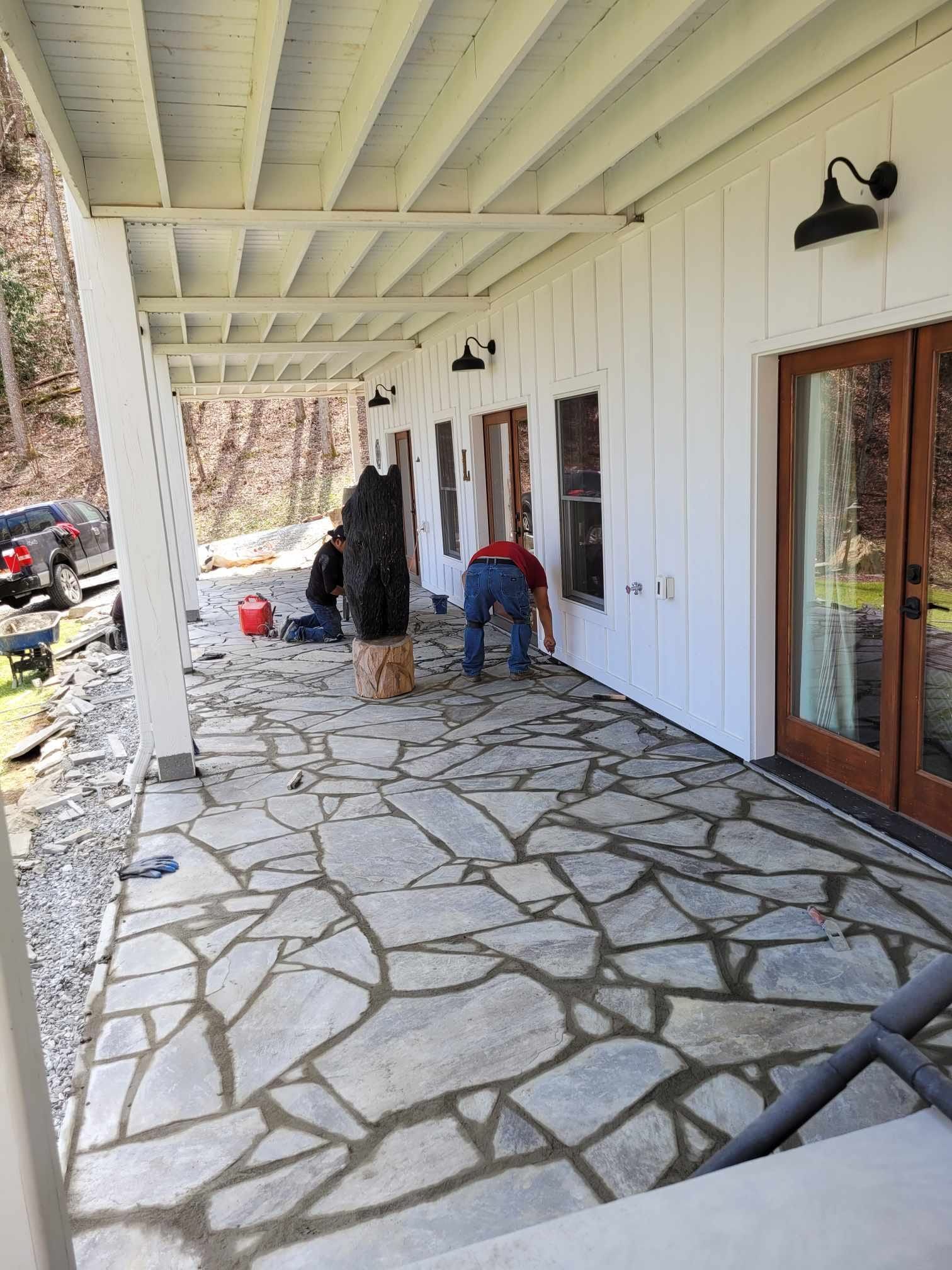 People work on a stone patio of a white house with brown doors, under a covered porch with a view of a wooded area.