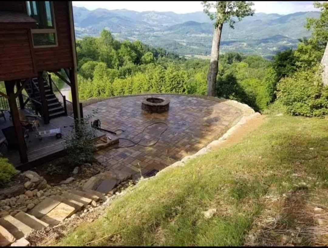 A stone patio with a fire pit overlooks a green valley.  Steps lead down a grassy hillside. A house is to the left.