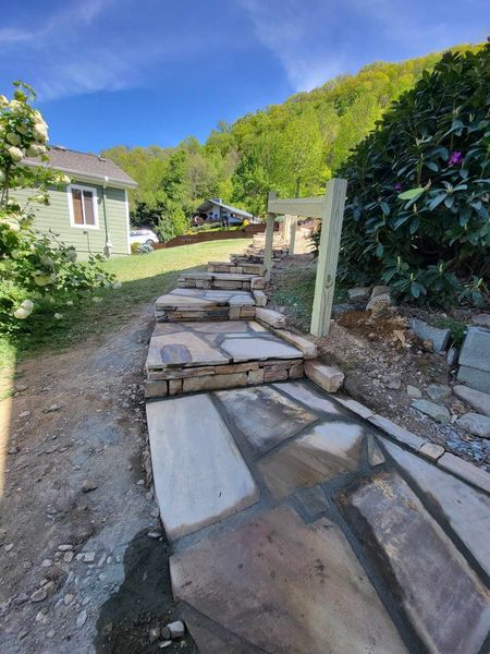 Stone steps lead up a hillside. Flagstone pathway and new wooden posts visible. Green trees and house in the background.